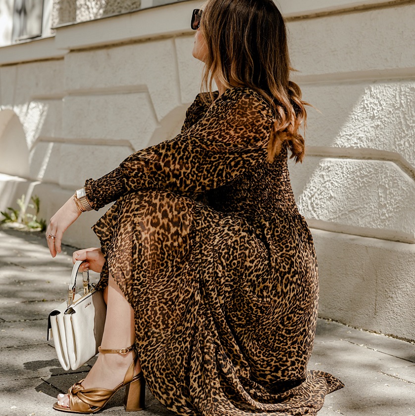picture of a woman standing on a sidewalk wearing dress with leopard print and brown sandals holding white bag