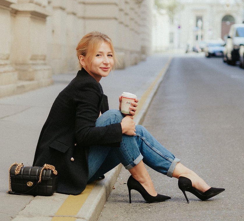 picture of a woman sitting on the sidewalk with a cup of coffee in her hand, wearing coat jeans and black sandals 