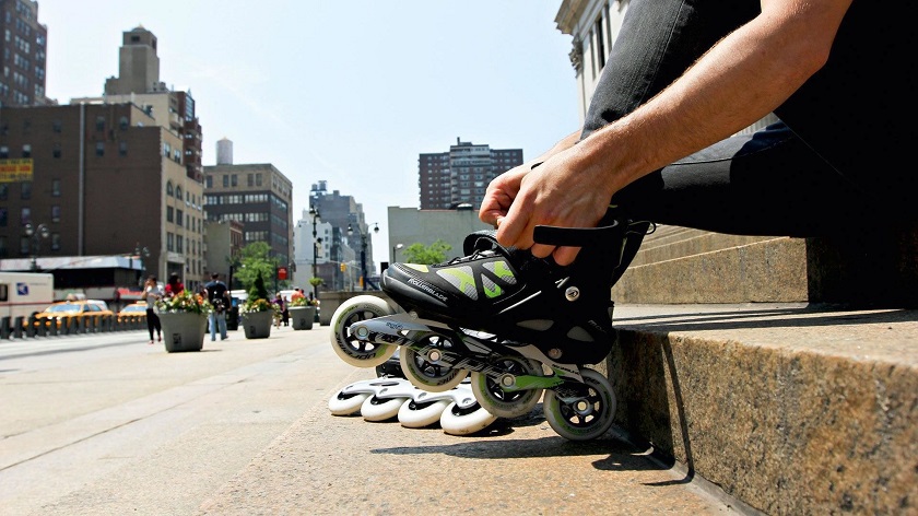 picture of a person putting his inline skates on while sitting on a concrete beside buildings 